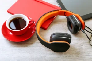 A cup of coffee in a red cup next to orange headphones and a tablet on a wooden surface.