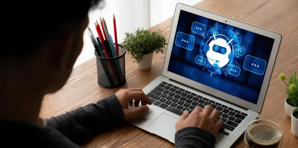 A person using a laptop displaying an AI and chatbot concept on screen, with a cup of coffee and office supplies on the table.
