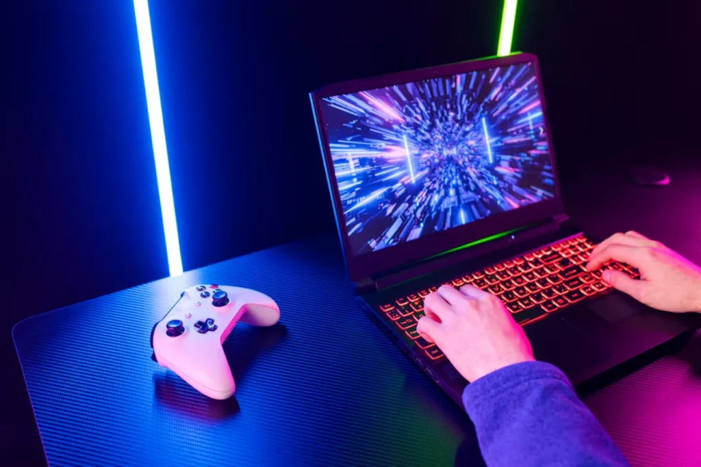 A person plays on a gaming laptop displaying vivid hyperspace-like graphics, with a white video game controller nearby, under neon blue lighting.