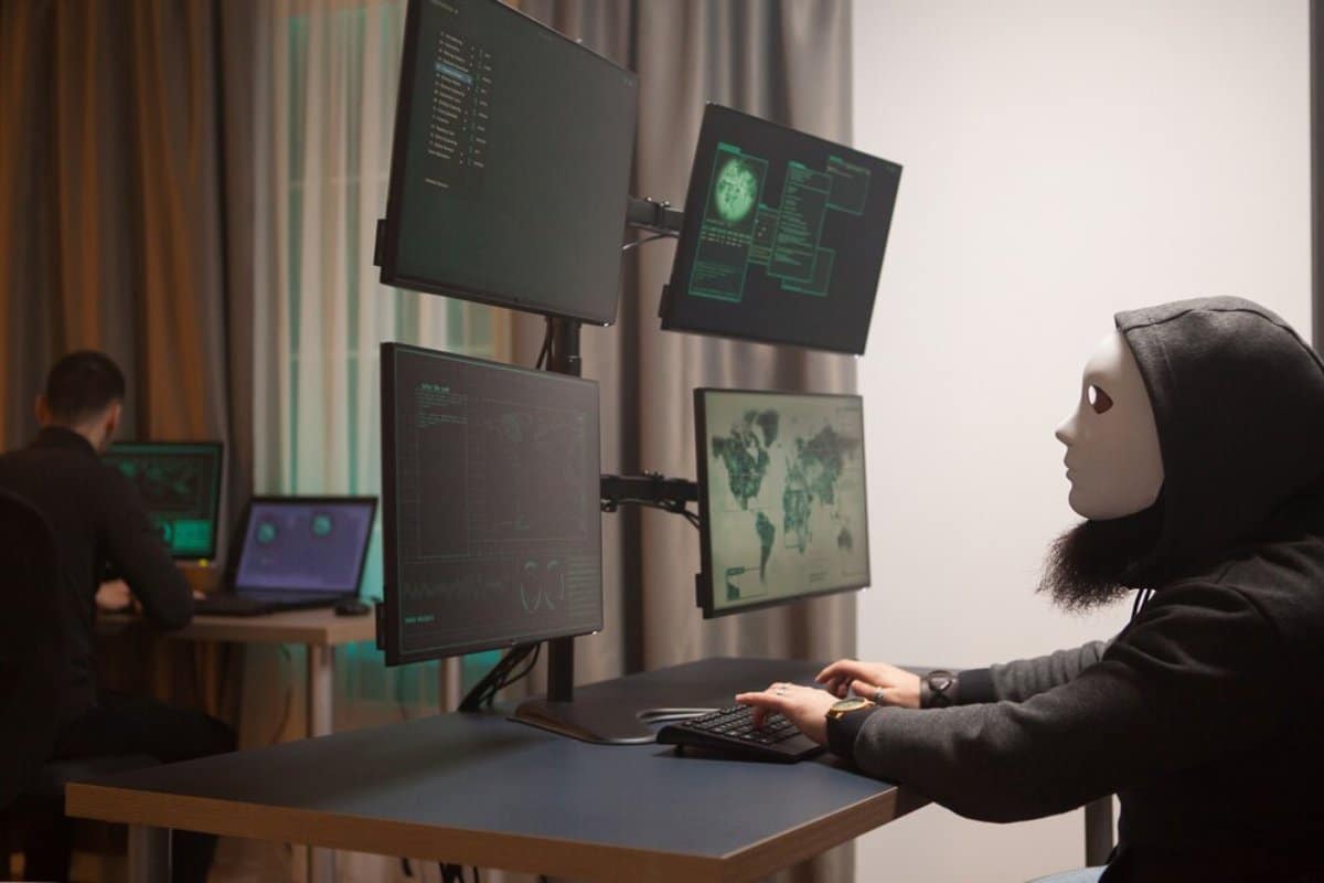 Two people, one in a mask, working at computer stations with multiple monitors displaying data and maps.