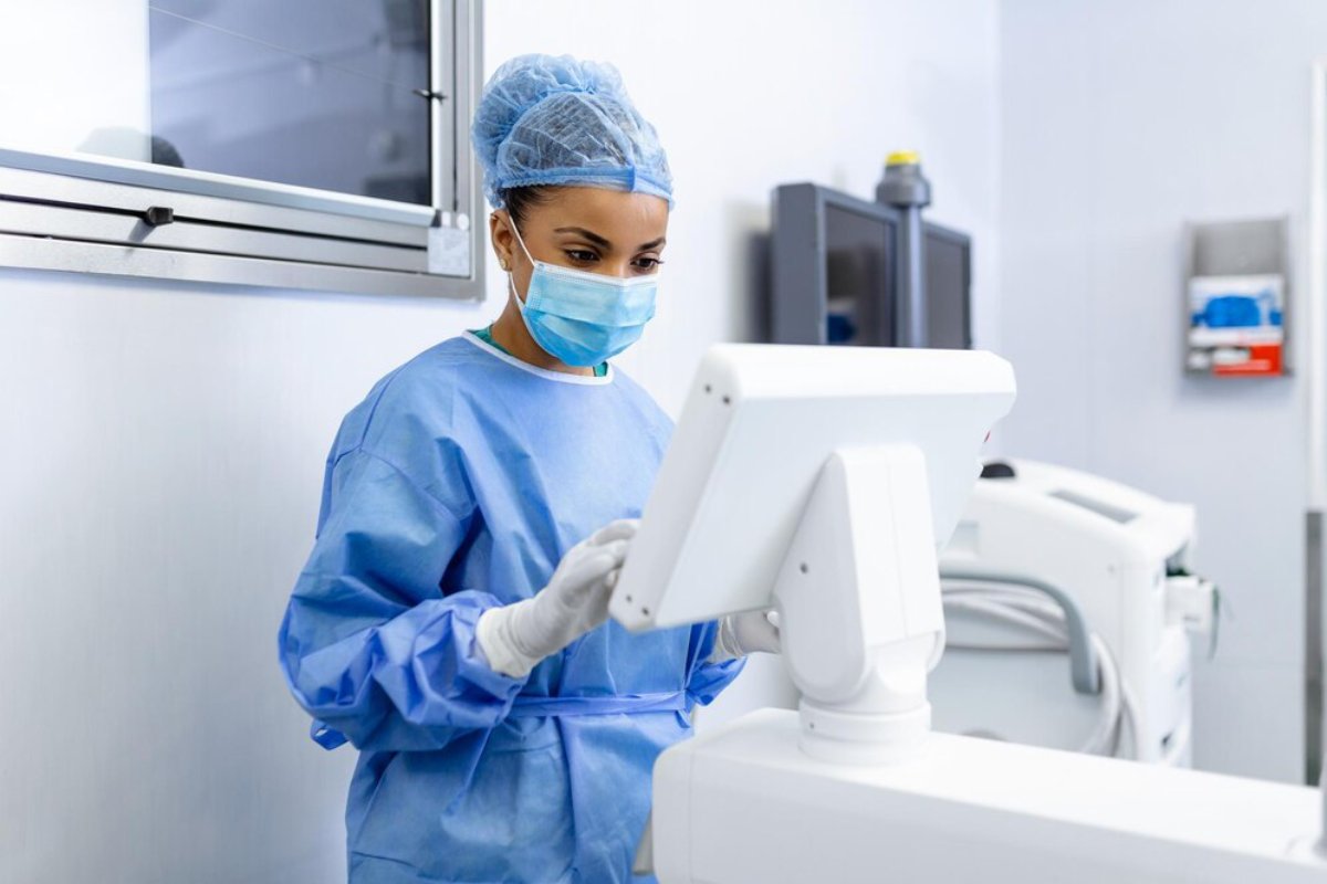 A healthcare professional in scrubs and gloves operates a medical monitor in a clean, well-lit healthcare setting.