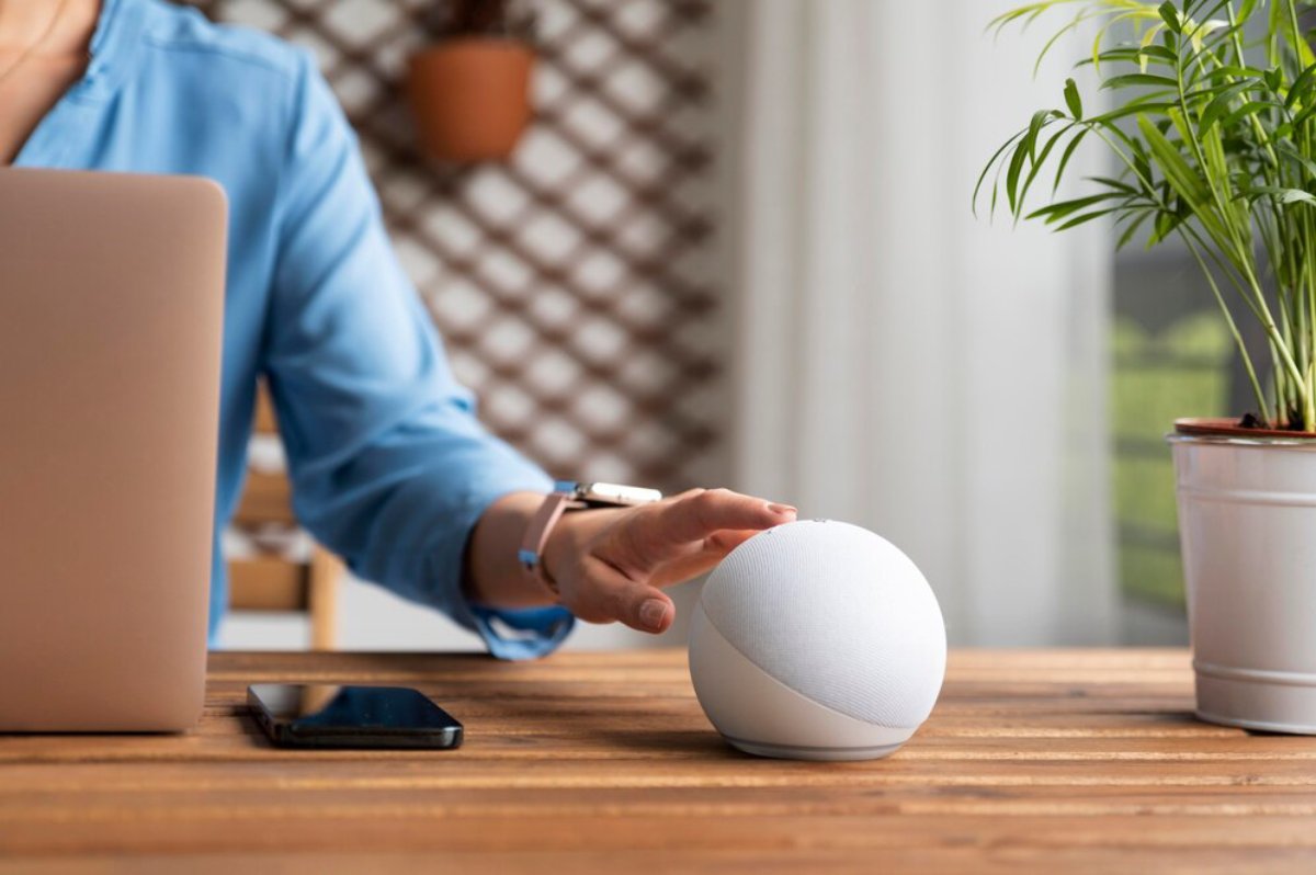 A person in a blue shirt interacts with a white smart speaker on a wooden desk, with a laptop and phone nearby, and a plant in a pot.