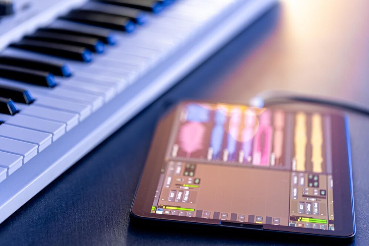 A close-up of a keyboard and a tablet displaying music editing software, connected by a cable on a dark surface.