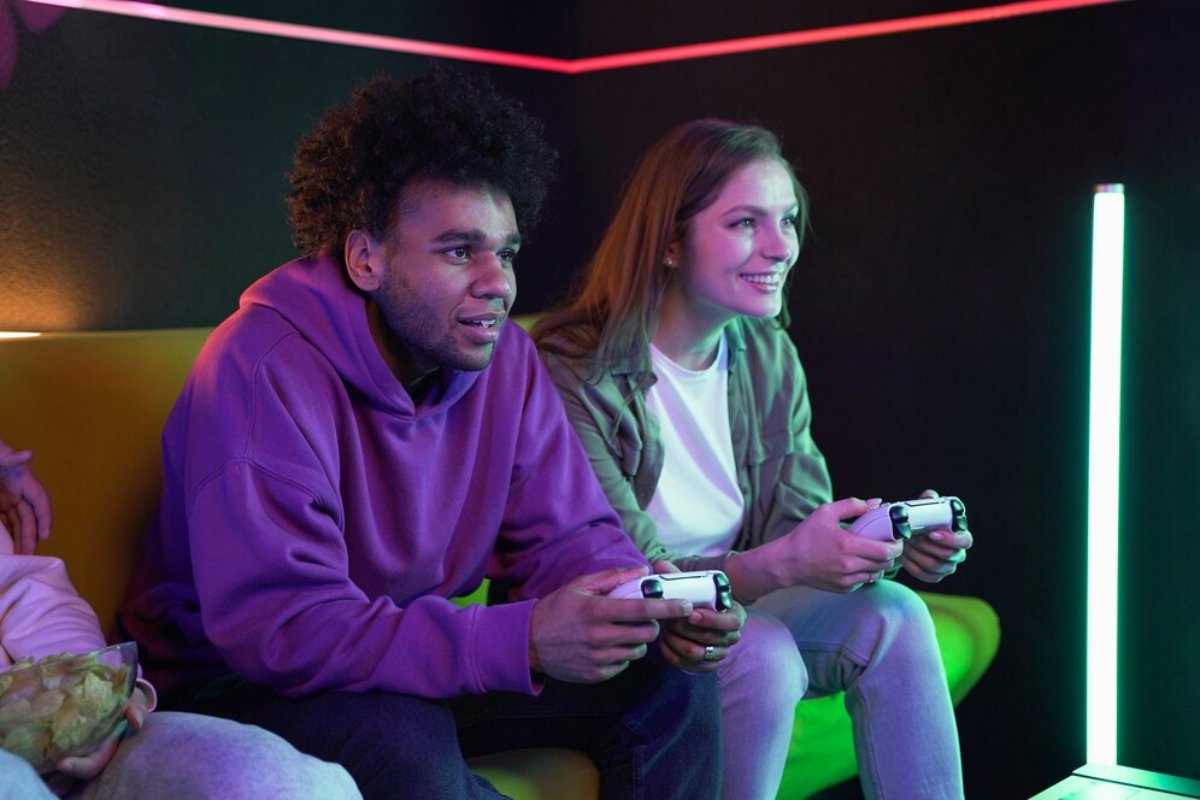 Two gamers sit on a colorful couch, focused on their game controllers amid glowing neon lights in a cozy gaming space.