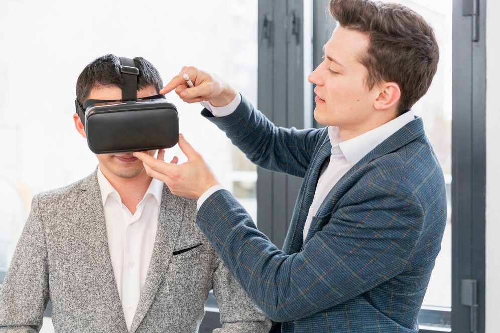 A man in a gray blazer adjusts a headset on another man wearing a light suit, both indoors near large windows.