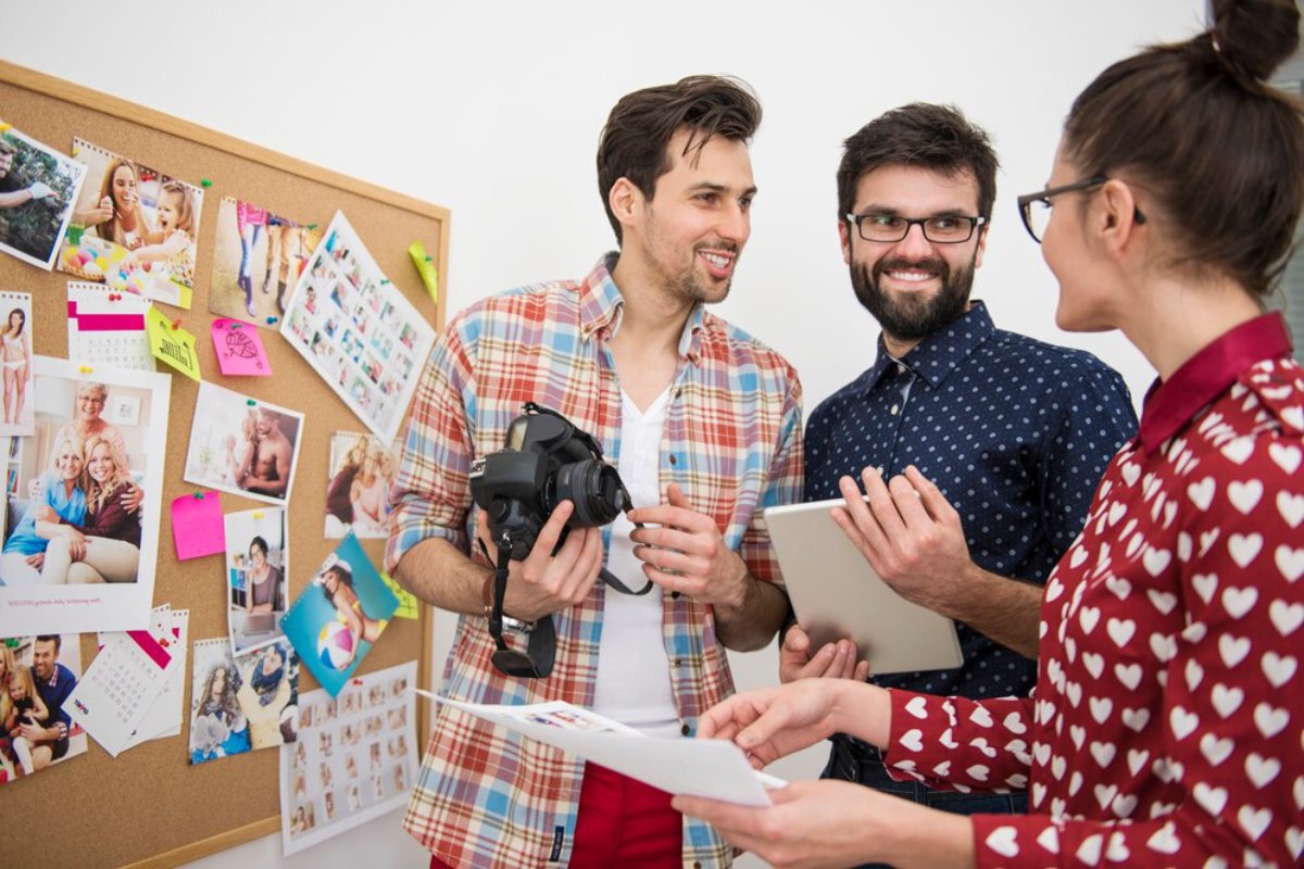 A group of three people discuss photography projects in front of a bulletin board filled with photos and sticky notes.
