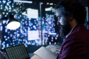 Man with a beard working at night, analyzing data on multiple computer screens in a dimly lit office.