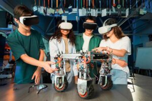 A group of four students collaborating on a robot with wheels, surrounded by various tech and tools in a workshop setting.