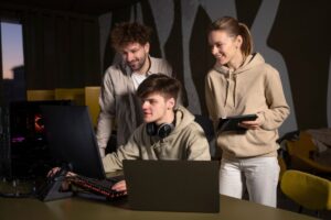 A group of three individuals collaborate at a desk with computers and a tablet, immersed in a tech-focused environment.