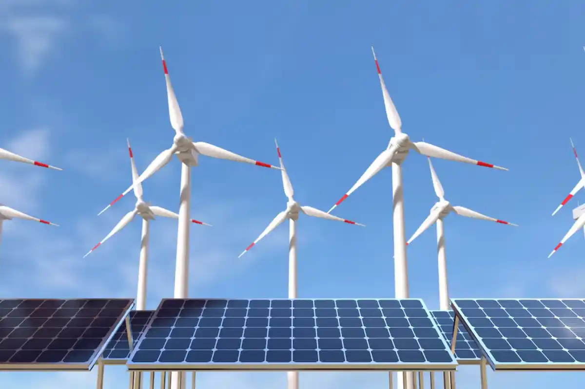 A row of photovoltaic solar panels in the foreground with multiple wind turbines against a clear blue sky in the background, symbolizing renewable energy sources.