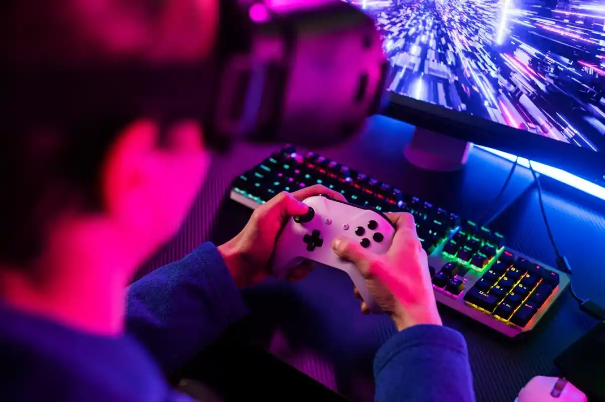 Person wearing a VR headset, holding a game controller while playing a fast-paced game on a monitor, with a colorful RGB-lit keyboard on the desk.