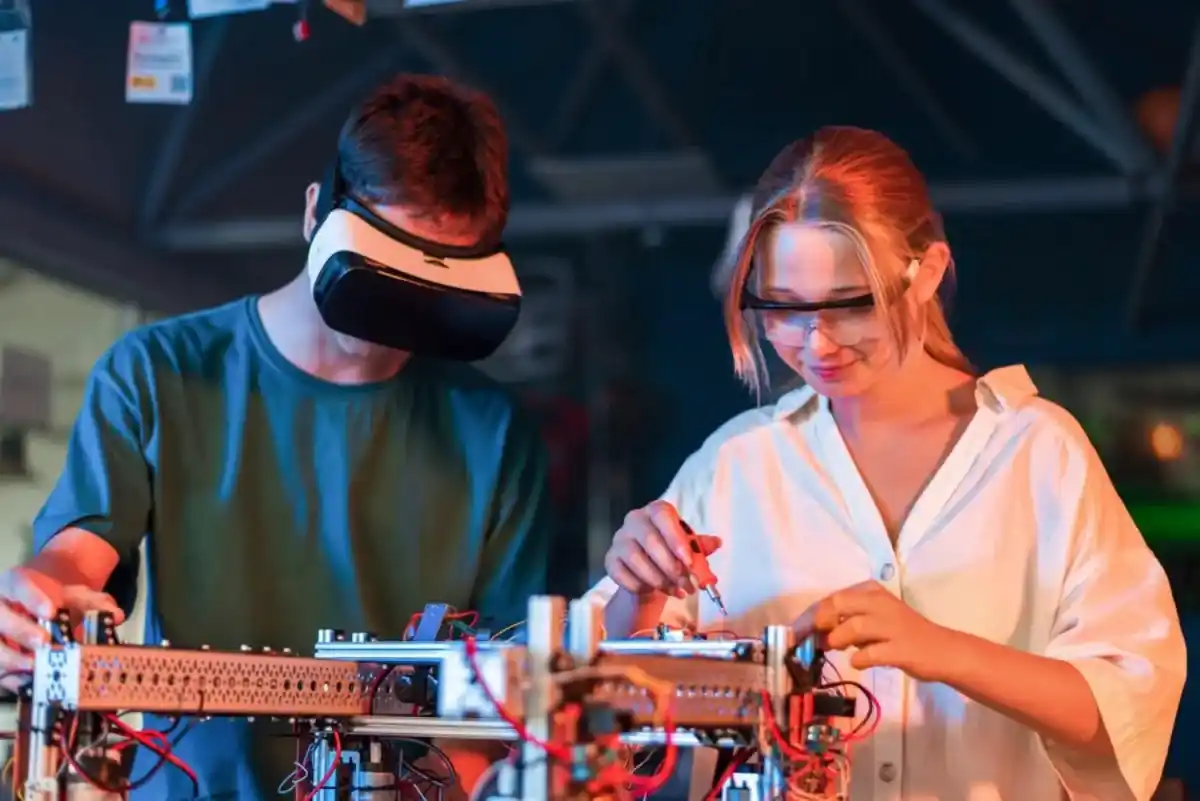 Two young engineers, a man wearing a VR headset and a woman with safety glasses, working collaboratively on an electronic robot project in a laboratory setting.