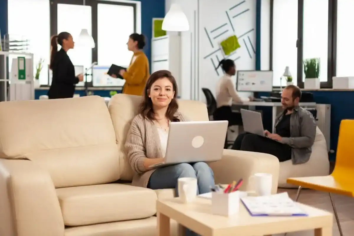 A young woman sitting comfortably on a cream-coloured sofa in a modern office setting, working on her laptop with colleagues engaging in work around her in the background.