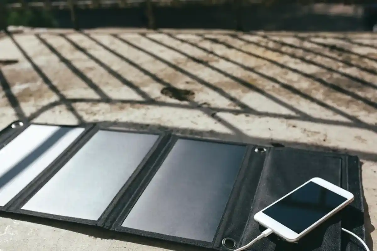A portable solar panel charger lying on a concrete surface with strong shadow lines, connected via cable to a smartphone.