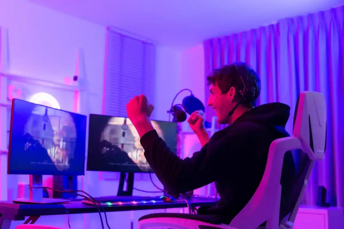 Young man wearing a headset, sitting at a gaming setup with two monitors, celebrating while playing a video game in a room lit with vibrant purple and blue lights.